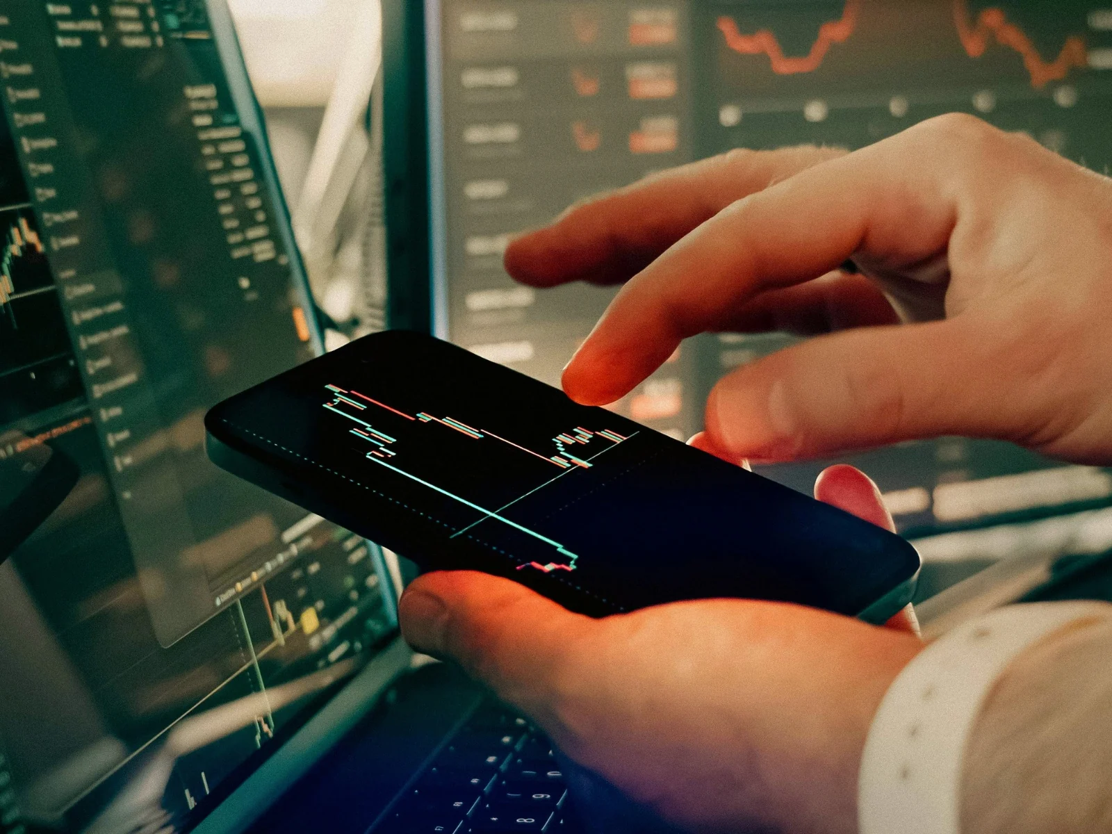 Persona consultando gráficos de inversión en un teléfono y varias pantallas